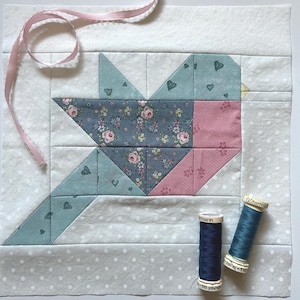 Op de afbeelding: Een stoffen vogel quiltblok met een roze lint en twee klosjes garen. De vogel is gemaakt van blauwe en roze stof met patronen. De achtergrond is wit met een grijze stippelrand.