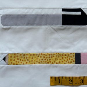 以下が含まれることがあります： 白い布地に、鉛筆の布製アップリケが2つ付いている。上の鉛筆はグレーで、黒い消しゴムが付いている。下の鉛筆は黄色で、黒とピンクのアクセントが付いている。画像の下部には、黒い目盛りが付いた黄色の定規がある。