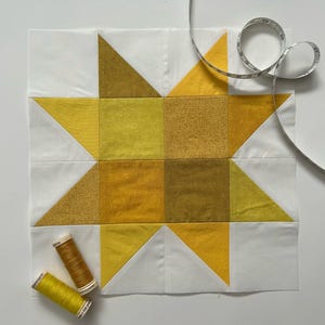 May include: A handmade quilt block featuring a star design in shades of yellow and white. The block is accompanied by two spools of thread and a measuring tape. The quilt block is approximately 10 inches square.