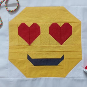 Puede incluir: Un cuadrado de tela amarillo con una cara sonriente hecha de corazones rojos para los ojos y un bigote negro. El cuadrado de tela está rodeado por un borde de tela blanco.