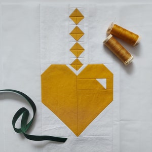 May include: A white fabric square with a yellow heart and a string of yellow diamonds at the top. Two spools of yellow thread are on the right side of the image. A green ribbon is on the left side of the image.