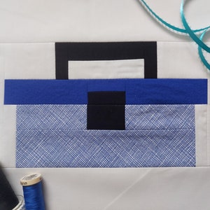 May include: A patchwork quilt block in the shape of a toolbox. The block is made of white, black, and blue fabric. The toolbox has a handle and a small compartment on the top.