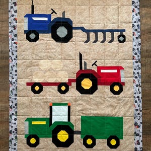 Może przedstawiać: Kołdra przedstawiająca trzy różne traktory. Górny traktor jest niebieski z pługiem, środkowy traktor jest czerwony z belą siana, a dolny traktor jest zielony z wozem.