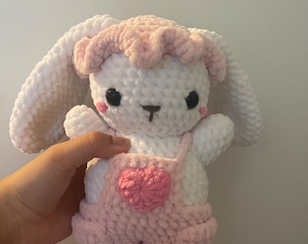 May include: A white crocheted bunny with pink ears and a pink crocheted hat. The bunny is wearing pink overalls with a pink heart on the front.