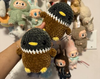 Puede incluir: Dos peluches de pingüino de crochet marrón y negro con picos amarillos y detalles blancos. Los peluches se sostienen en una mano contra una pared blanca con otros peluches en el fondo.