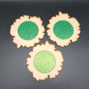 May include: Three wooden coasters with a unique design. Each coaster has a light brown, irregular outer shape with a textured green circle in the center. The coasters are arranged on a dark background.