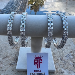 May include: Four silver bracelets with clear cubic zirconia stones. The bracelets are stacked on a white stand with a logo that says "Rida Fashionz".