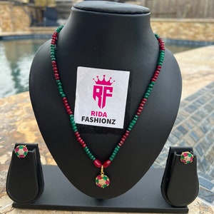 May include: A green and red beaded necklace with a gold pendant featuring a green and red stone surrounded by clear stones. The necklace is displayed on a black stand with matching earrings.