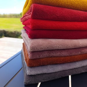 Puede incluir: Una pila vertical de siete toallas rectangulares dobladas en una variedad de colores que incluyen amarillo mostaza, rojo brillante, carmesí, rosa viejo, malva apagado, naranja quemado y gris claro, todas con una superficie de tela texturizada.