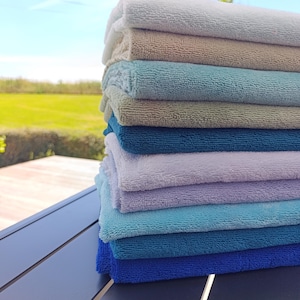 Puede incluir: Una pila vertical de diez paños de microfibra rectangulares doblados en un degradado de colores que incluye blanco, beige, verde claro, verde azulado, gris claro, lavanda, azul claro y azul oscuro, con una superficie de tela texturizada.