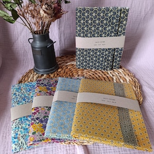 Può includere: Una collezione di tovaglioli di stoffa con motivi floreali e geometrici, tra cui blu, giallo e viola. Ogni tovagliolo è avvolto con una fascia di carta. A sinistra, un piccolo vaso di metallo scuro con fiori secchi.