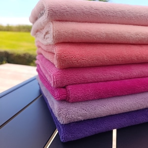 Puede incluir: Una pila de siete toallas rectangulares dobladas dispuestas en una secuencia de colores degradados. Los tonos transicionan desde un rosa pálido en la parte superior, pasando por matices coral, rosa y magenta, hasta un morado intenso.