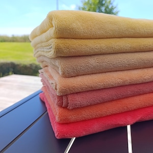 Puede incluir: Una pila de seis toallas de microfibra dobladas dispuestas en un patrón de degradado de color, que va desde un amarillo pálido en la parte superior hasta un rosa intenso vibrante en la parte inferior. Cada toalla tiene una superficie de tela suave.