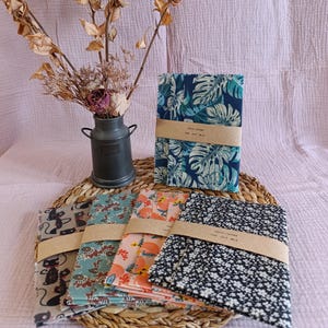 Peut inclure: Plusieurs emballages alimentaires en tissu avec différents motifs : feuilles tropicales bleues, motifs floraux et chats. Chaque emballage est maintenu par une bande de papier marron. Un vase en métal en forme de pot à lait avec des fleurs séchées est également présent.