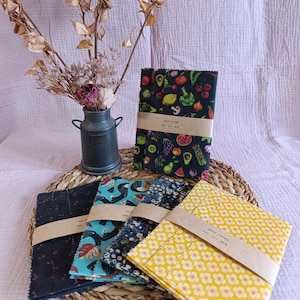 Può includere: Una collezione di involucri alimentari riutilizzabili con vari motivi, tra cui frutta, fiori e disegni astratti. Gli involucri sono raggruppati con fasce di carta marrone. È presente anche un piccolo vaso di metallo con fiori secchi.