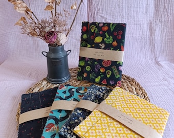 May include: A collection of reusable food wraps with various patterns, including fruits, flowers, and abstract designs. The wraps are bundled with brown paper bands. A small metal vase with dried flowers sits nearby on a woven mat.