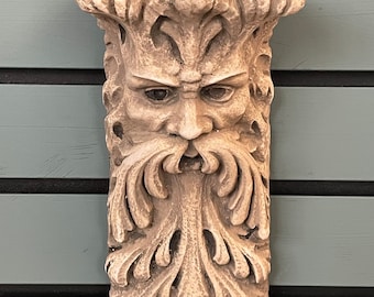 May include: A beige stone sculpture of a green man's face with a leafy beard. The sculpture is mounted on a wall and has a shelf on top.