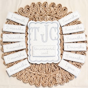 May include: A baby milestone photo prop set. The set includes a central, scalloped-edged plaque with the initials "TJC" and the name "townesend carl", surrounded by rectangular labels marking months 1-11 and 1 year. The items are light blue on a woven, beige background.