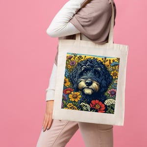 May include: A white canvas tote bag featuring a colourful illustration of a black dog surrounded by flowers. The dog is looking directly at the viewer.