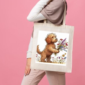 May include: A cream-coloured tote bag with a cartoon illustration of a brown dog holding a bouquet of colourful flowers. The dog is in a field of wildflowers. The background is white.