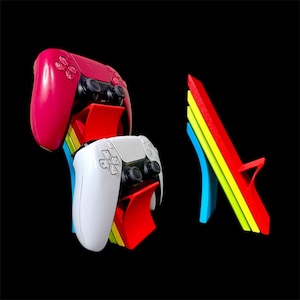 Pode incluir: Uma imagem vibrante mostra dois controles de jogos, um vermelho e um branco, apoiados em suportes coloridos. Os suportes são um arco-íris de cores, incluindo vermelho, amarelo e azul. O fundo é preto, destacando os controles e os suportes.