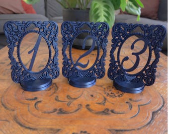May include: Three navy blue table number holders. Each holder has an oval frame with ornate detailing and a number from one to three. The holders are on a dark brown wooden table.