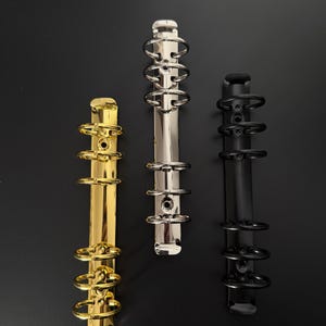Könnte beinhalten: Drei Metall-Ringbuchmechaniken mit jeweils sechs Ringen, vertikal angeordnet. Die linke Mechanik ist goldfarben, die mittlere silberfarben und die rechte mattschwarz. Jede verfügt über eine zentrale Schiene mit kreisförmigen Ringen.