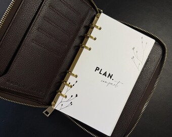 Könnte beinhalten: Ein brauner Leder-Reißverschluss-Planer mit goldenen Ringen und einer weißen Seite, die auf eine Seite mit der Aufschrift "PLAN. compact." geöffnet ist.