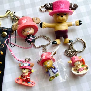 May include: A collection of colorful anime character keychains. The keychains feature a cartoon character with a pink hat and antlers in various poses, including one on a snowboard and another in a bowl. The keychains are made of plastic.