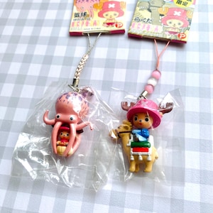 May include: Two anime-themed mobile phone charms in clear plastic packaging. One charm features a small character inside a pink octopus, and the other shows a character riding a tan camel. Both include colorful header cards with Japanese text.