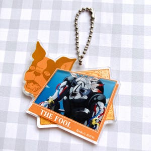 May include: A double-layered acrylic keychain featuring a Boston Terrier head in orange and a rectangular card design labeled THE FOOL. The card depicts a mechanical, dark-colored figure with white accents and red cables, attached to a silver ball chain.