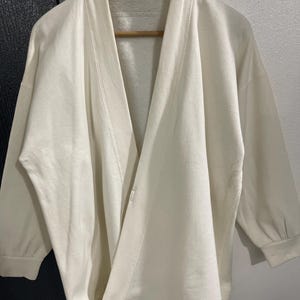 Pu&ograve; includere: Accappatoio color crema con collo a scialle e maniche a tre quarti. L'accappatoio &egrave; realizzato in un materiale morbido ed &egrave; appeso a un'appendiabiti in legno. L'etichetta recita "robe de chambre".