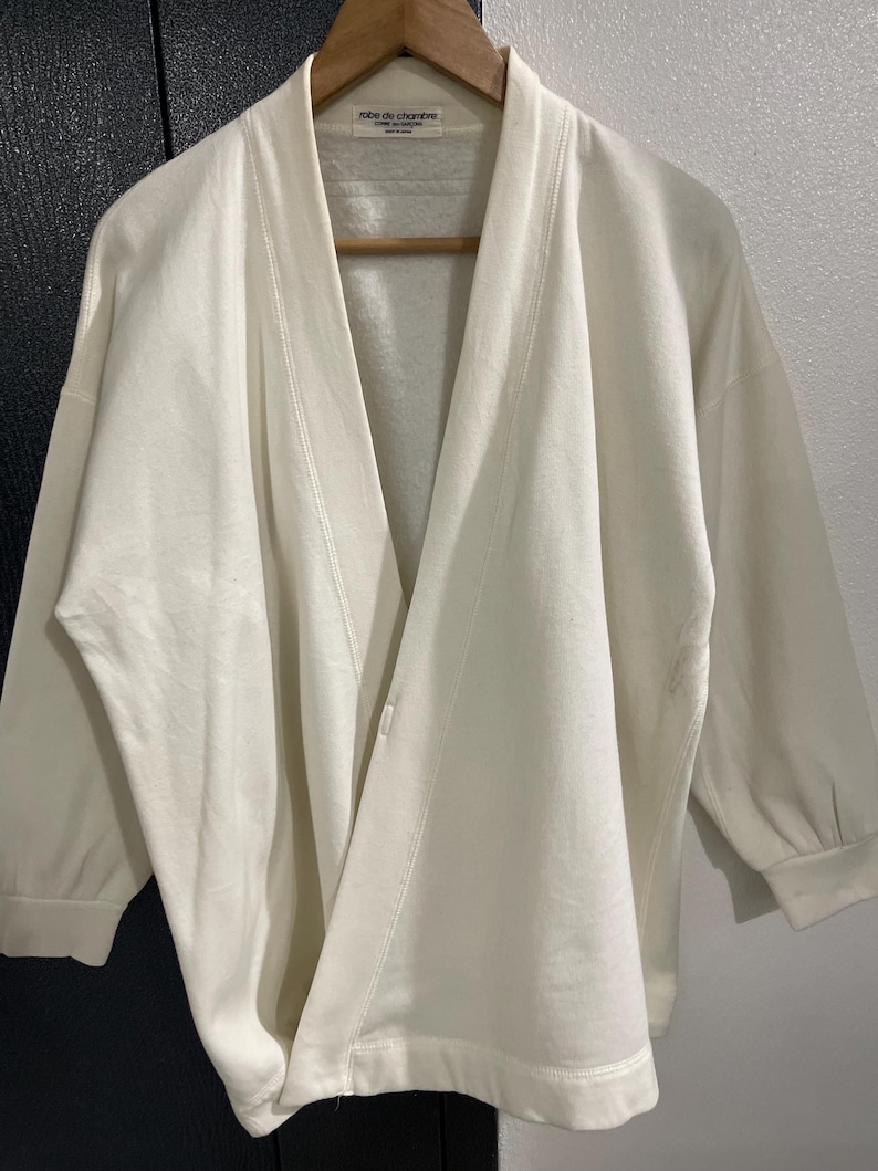 Pu&ograve; includere: Accappatoio color crema con collo a scialle e maniche a tre quarti. L'accappatoio &egrave; realizzato in un materiale morbido ed &egrave; appeso a un'appendiabiti in legno. L'etichetta recita "robe de chambre".