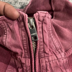 Peut inclure: Gros plan sur une veste rose poussi&eacute;reux avec une fermeture &eacute;clair argent&eacute;e. Le curseur de la fermeture &eacute;clair porte le nom de la marque "AVIREX". La veste a un col c&ocirc;tel&eacute; et un tissu textur&eacute;.