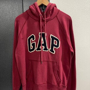 Może przedstawiać: Bordowa bluza z kapturem z napisem "GAP" w dużych granatowych literach z białym obramowaniem na piersi. Bluza ma kaptur ze sznurkiem i przednią kieszeń. Ubranie wisi na drewnianym wieszaku.