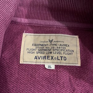 Peut inclure: Gros plan d'un sweat &agrave; capuche bordeaux avec une &eacute;tiquette indiquant "EQUIPMENT-TYPE-AVIREX" et "AVIREX LTD". L'&eacute;tiquette comprend &eacute;galement le texte "FLIGHT GARMENT SPECIFICATION" et "HIGH SPEED LOW LEVEL FLIGHT". Le sweat &agrave; capuche est de taille XL.