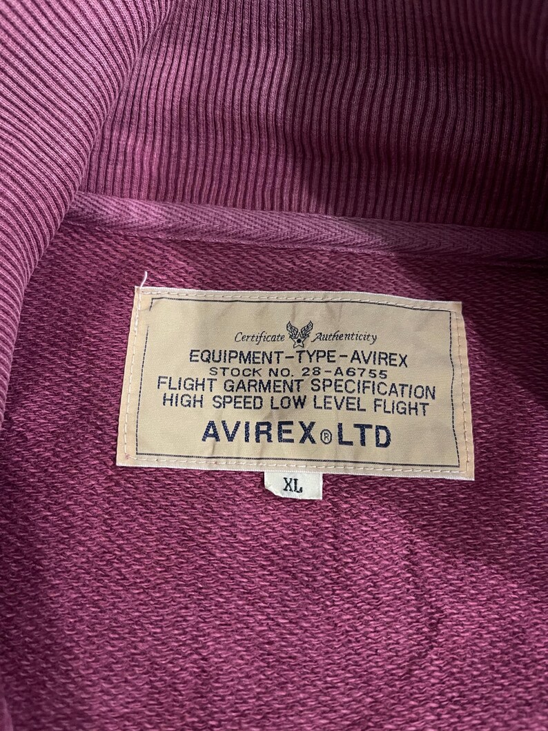 Peut inclure: Gros plan d'un sweat &agrave; capuche bordeaux avec une &eacute;tiquette indiquant "EQUIPMENT-TYPE-AVIREX" et "AVIREX LTD". L'&eacute;tiquette comprend &eacute;galement le texte "FLIGHT GARMENT SPECIFICATION" et "HIGH SPEED LOW LEVEL FLIGHT". Le sweat &agrave; capuche est de taille XL.