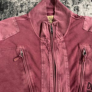 Peut inclure: Gros plan sur une veste zipp&eacute;e rose poussi&eacute;reux. La veste a un col c&ocirc;tel&eacute;, deux poches zipp&eacute;es et une fermeture &eacute;clair pleine longueur. La veste a un aspect vintage d&eacute;lav&eacute;. Le texte "DU" et "USA A" est visible &agrave; droite.