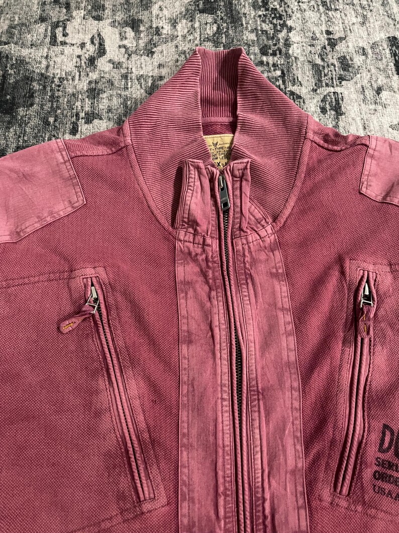 Peut inclure: Gros plan sur une veste zipp&eacute;e rose poussi&eacute;reux. La veste a un col c&ocirc;tel&eacute;, deux poches zipp&eacute;es et une fermeture &eacute;clair pleine longueur. La veste a un aspect vintage d&eacute;lav&eacute;. Le texte "DU" et "USA A" est visible &agrave; droite.
