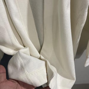 Pu&ograve; includere: Primo piano di una felpa color crema. Il tessuto appare morbido e spesso, con cuciture visibili lungo le cuciture e i polsini. I polsini sono ripiegati e il design generale suggerisce un capo casual e confortevole.