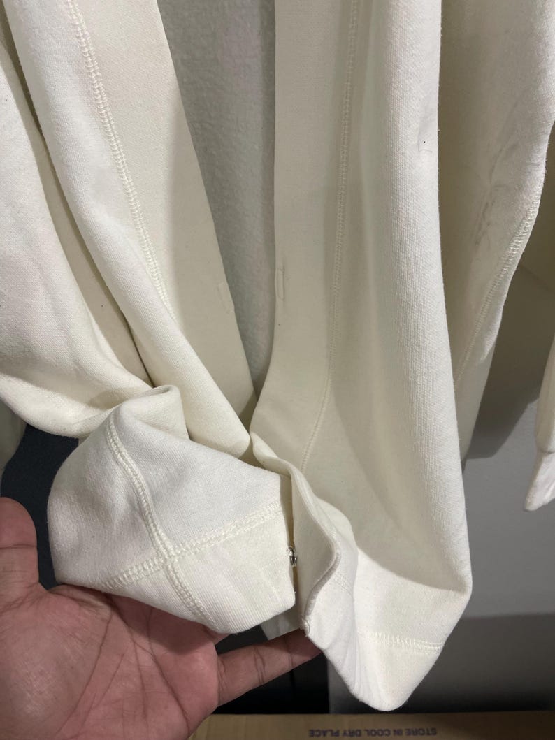 Pu&ograve; includere: Primo piano di una felpa color crema. Il tessuto appare morbido e spesso, con cuciture visibili lungo le cuciture e i polsini. I polsini sono ripiegati e il design generale suggerisce un capo casual e confortevole.