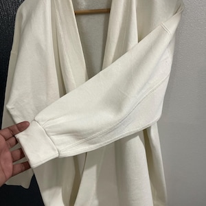 Pu&ograve; includere: Felpa robe de chambre color crema con collo a scialle e maniche lunghe. L'indumento &egrave; realizzato in un materiale morbido e spesso e presenta una vestibilit&agrave; ampia. Il testo "robe de chambre" &egrave; visibile sull'etichetta.