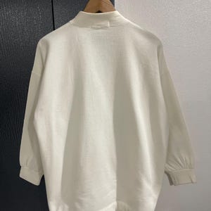 Pu&ograve; includere: Felpa color crema con collo alto e maniche lunghe. Le maniche hanno un dettaglio arricciato al polso. Il capo &egrave; appeso a una gruccia di legno su uno sfondo neutro.