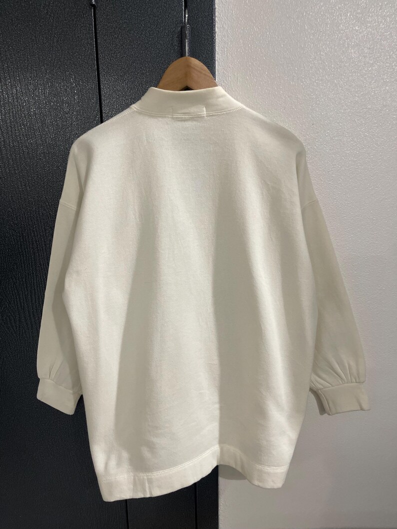 Pu&ograve; includere: Felpa color crema con collo alto e maniche lunghe. Le maniche hanno un dettaglio arricciato al polso. Il capo &egrave; appeso a una gruccia di legno su uno sfondo neutro.