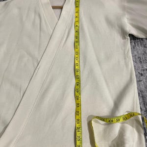 Pu&ograve; includere: Top avvolgente color crema con scollo a V e maniche lunghe. Un metro a nastro giallo &egrave; appoggiato sul capo, mostrando le misure in centimetri. Il top &egrave; realizzato in un materiale morbido, probabilmente cotone.