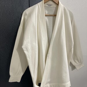 Pu&ograve; includere: Cardigan color crema con davanti aperto e maniche lunghe, appeso a una gruccia di legno. Il cardigan ha una vestibilit&agrave; ampia e una trama sottile. L'etichetta recita "robe de chambre".