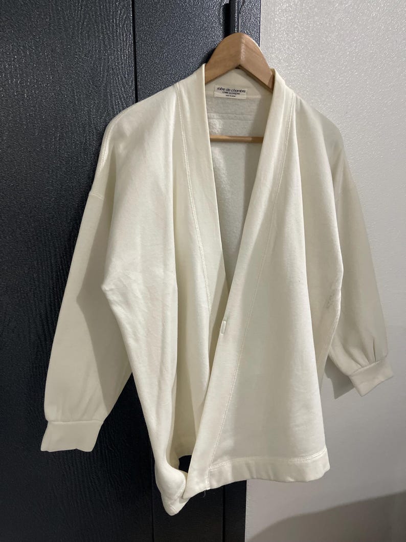 Pu&ograve; includere: Cardigan color crema con davanti aperto e maniche lunghe, appeso a una gruccia di legno. Il cardigan ha una vestibilit&agrave; ampia e una trama sottile. L'etichetta recita "robe de chambre".