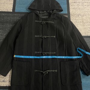 Może przedstawiać: Czarny wełniany płaszcz typu duffle coat z kapturem i zapięciami na kołki. Płaszcz posiada dwie przednie kieszenie i mierzy około 58 cm w poprzek klatki piersiowej. Płaszcz leży płasko na wzorzystej powierzchni.