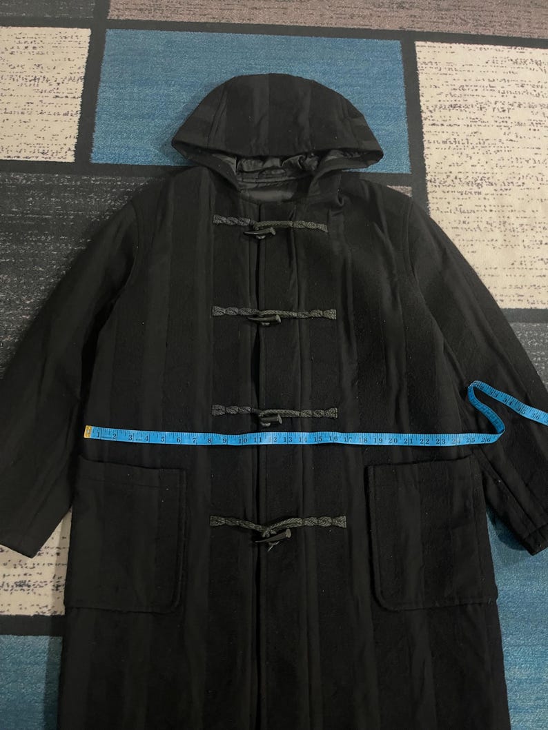 Może przedstawiać: Czarny wełniany płaszcz typu duffle coat z kapturem i zapięciami na kołki. Płaszcz posiada dwie przednie kieszenie i mierzy około 58 cm w poprzek klatki piersiowej. Płaszcz leży płasko na wzorzystej powierzchni.