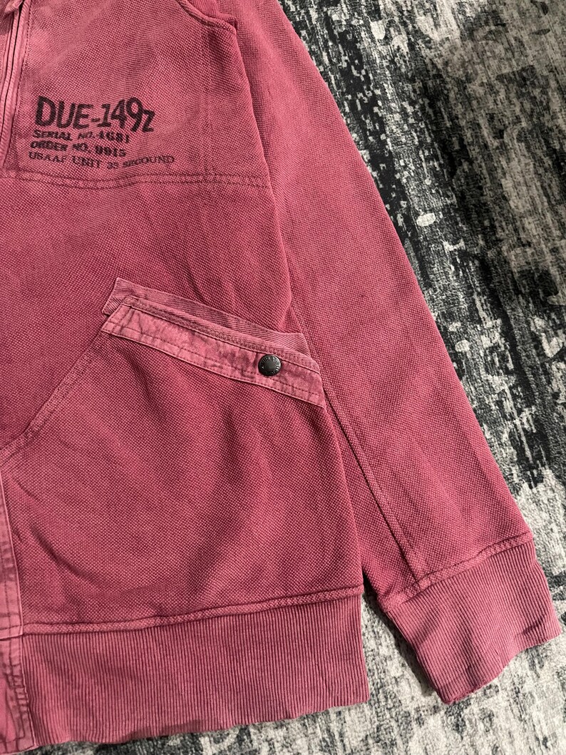 Peut inclure: Un sweat &agrave; capuche zipp&eacute; bordeaux avec une poche et des poignets c&ocirc;tel&eacute;s. La zone sup&eacute;rieure gauche de la poitrine porte le texte "DUE-1492" et d'autres textes. Le sweat &agrave; capuche est en tissu textur&eacute;.